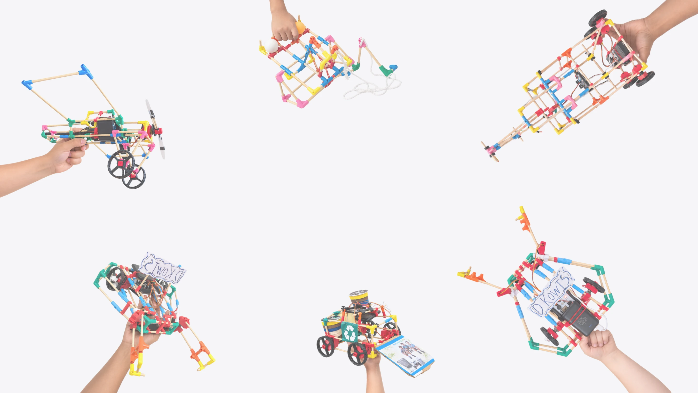 Six hands holding things students built from Stick ’Em: a propeller plane, a pulley rig, a buggy with a phone on it, a flag-waving cart and two machines with hand-drawn signs
