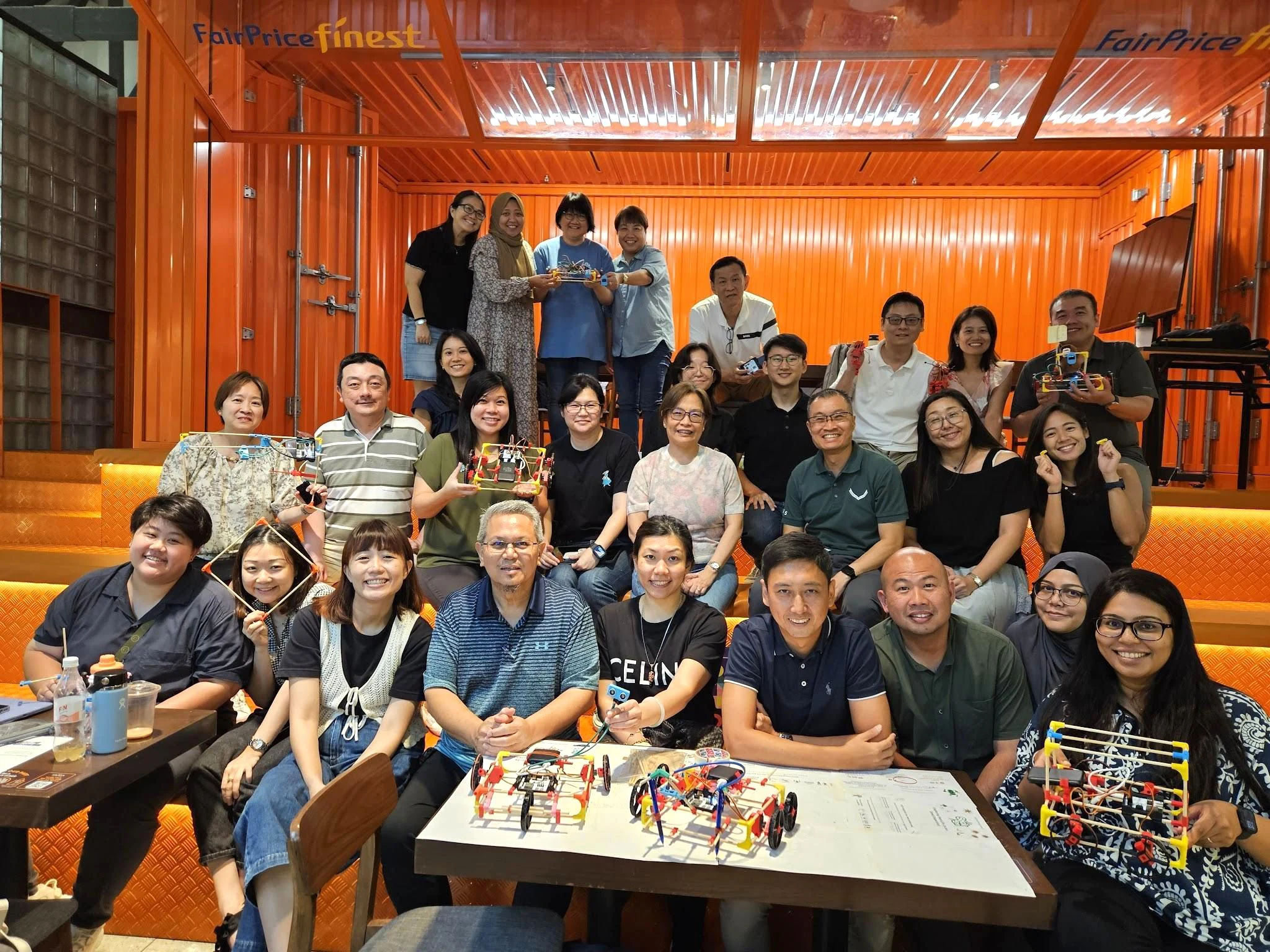 Teachers seated in a wood-panelled hall behind a table of the robots they built